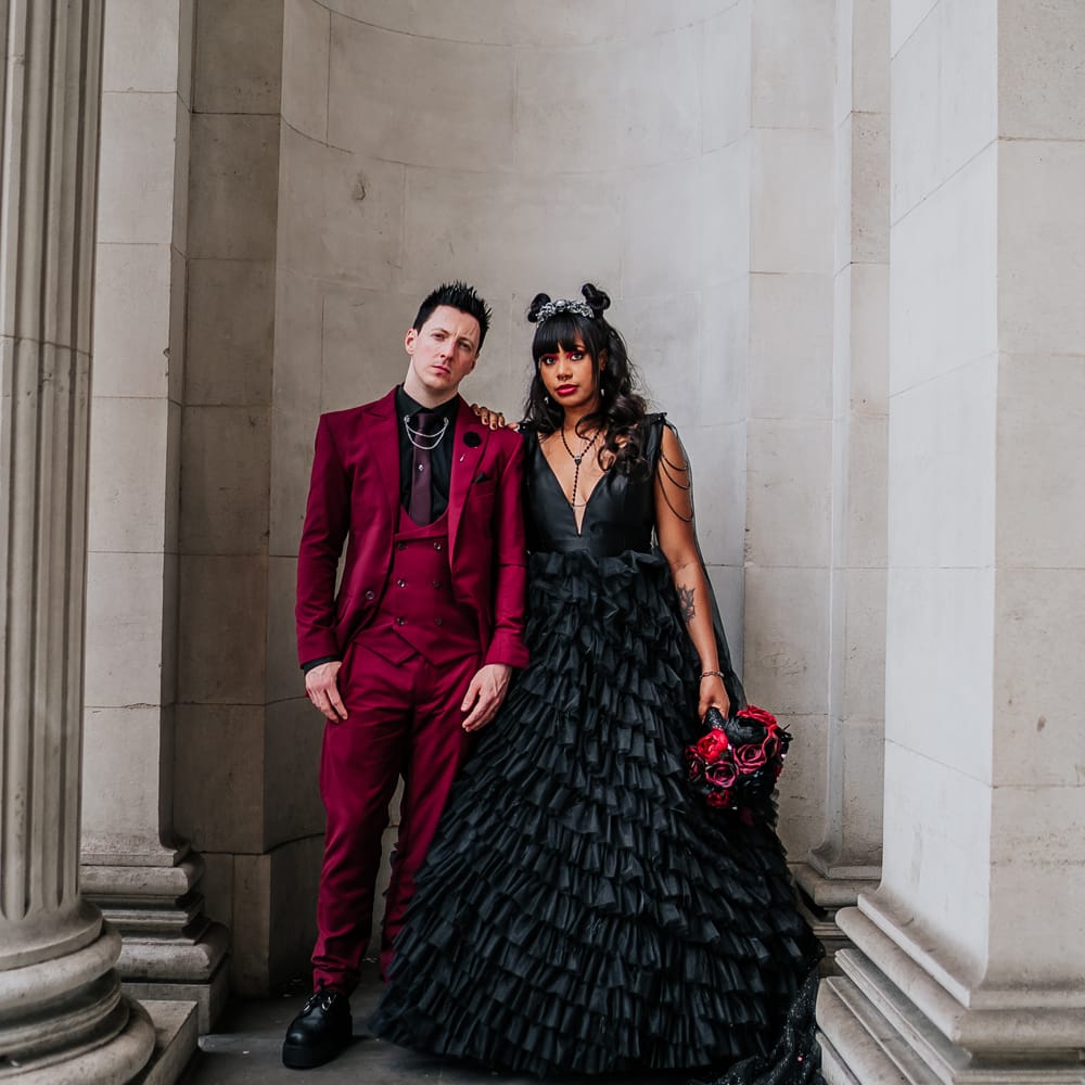 bride wearing black wedding dress stands beside her groom in wine red suit for gothic styled wedding day