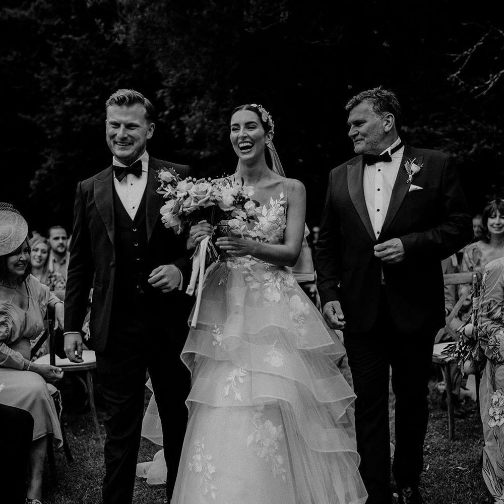 Bride in a layered tulle wedding dress standing with the groom and her father in black tie for the outdoor ceremony 