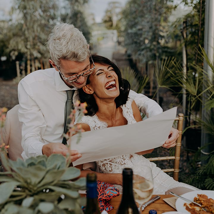 Laughing Bride leans on Groom who is reading while seated on rustic chairs