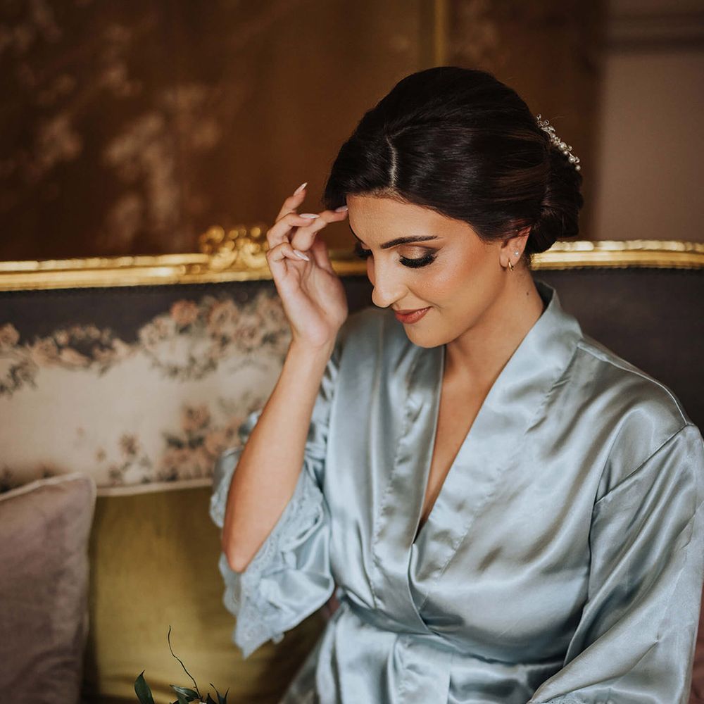 Bride wearing a blue satin robe with dark brown hair and smoky eye makeup holding a white flower bouquet 