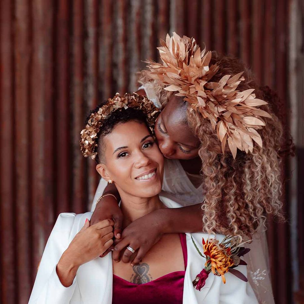 Bride in white silk suit with autumnal dried floral boutonniere and golden bridal crown being hugged by bride in large golden leaf bridal crown 