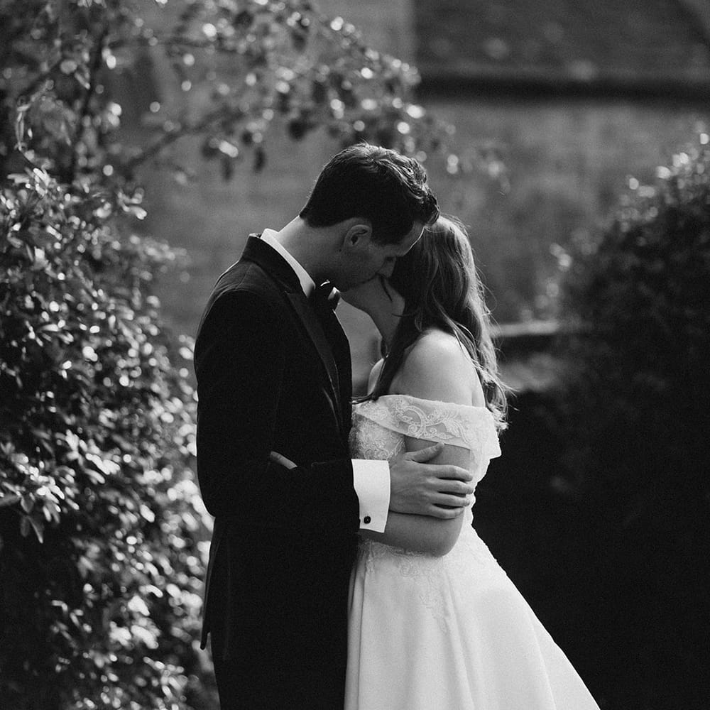 Groom in a tuxedo kisses the bride on the cheek in an off the shoulder Pronovias wedding dress for their traditional wedding 