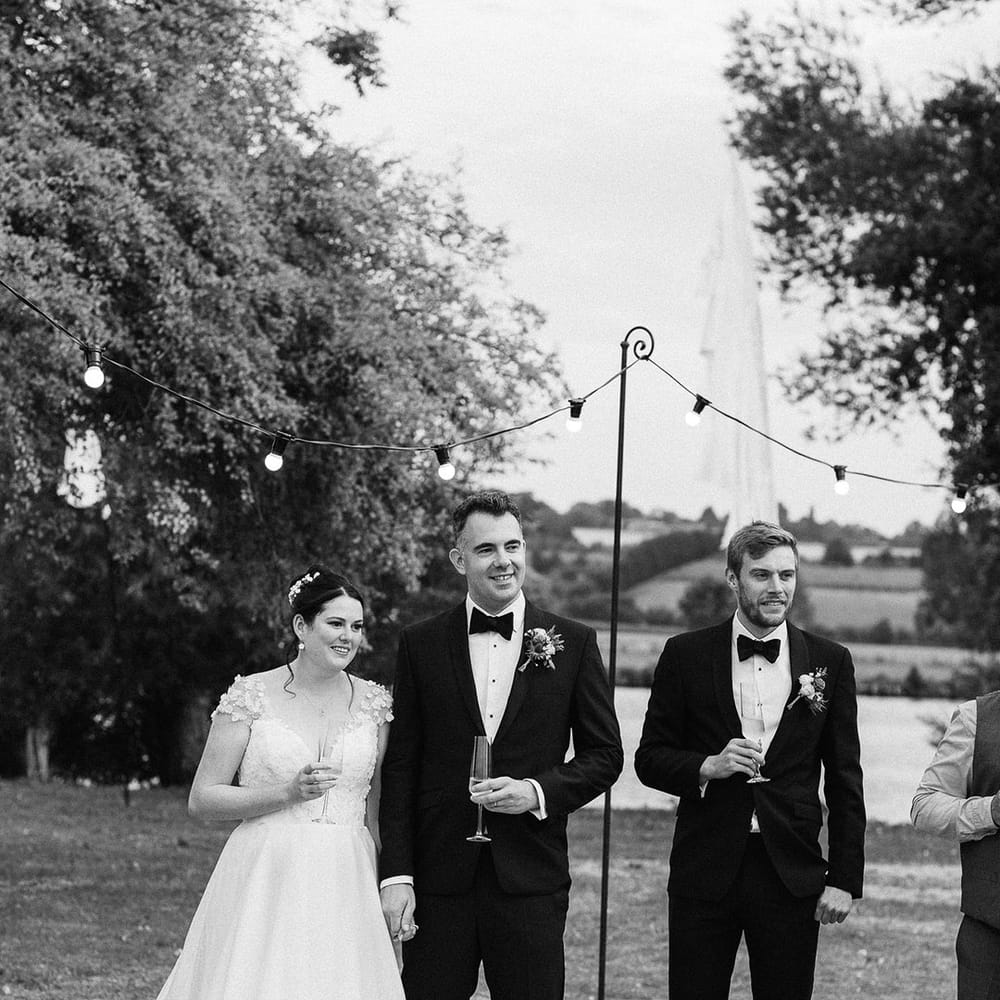 Festoon lighting decorates the outdoor area with the bride and groom standing together in their timeless wedding outfits sharing some wine 