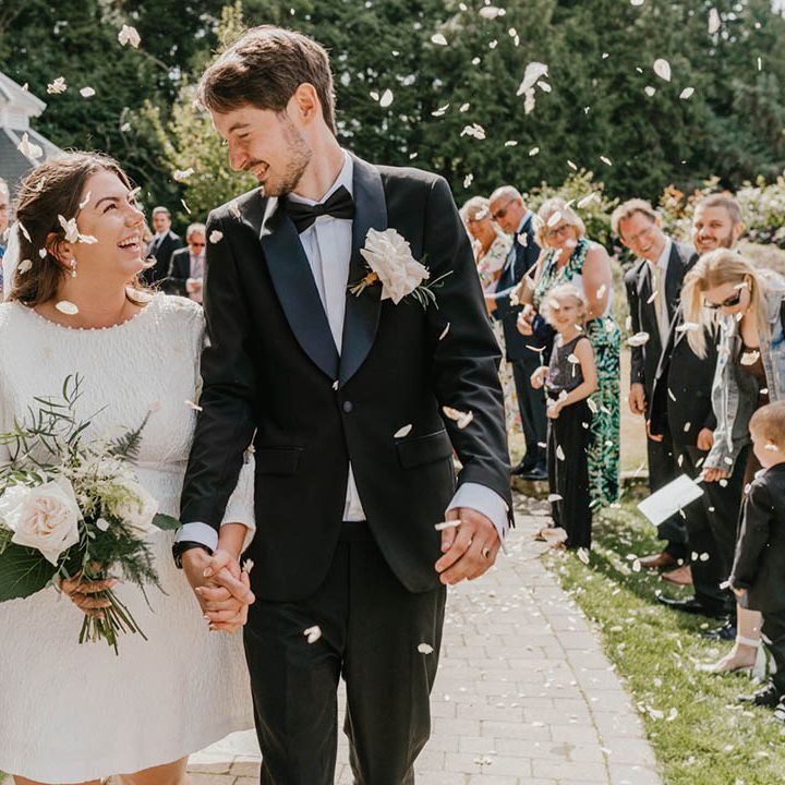 The bride and groom beam at each other on their wedding day with the bride in a short wedding dress with bow and groom in black tie as their guests throw confetti 