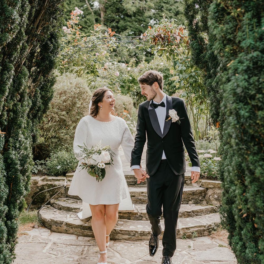 Groom in black tie with white rose buttonhole walking hand in hand with the bride in a long sleeve short wedding dress with a train and bow detailing 