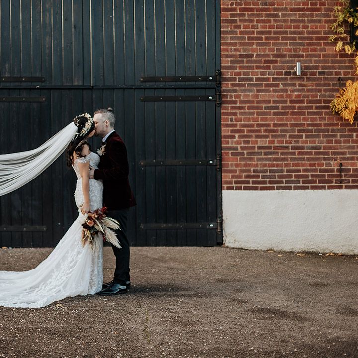 The groom in a burgundy velvet suit jacket kisses the bride in an off the shoulder lace wedding dress with the veil blowing in the wind 