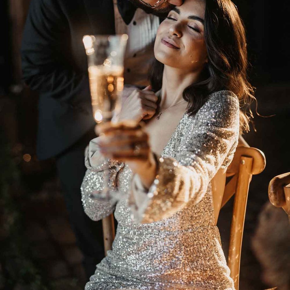 Bride in silver sparkly wedding dress holding glass of champagne, being kissed on the forehead by groom in classic black tuxedo and black bowtie 