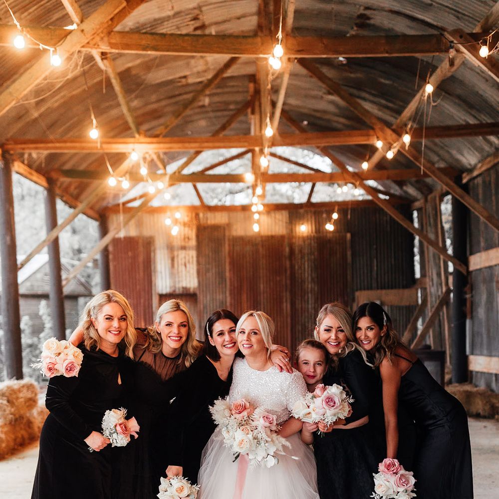 Bridesmaids in mismatched black bridesmaid dresses with the bride under the industrial shelter at Silchester Farm