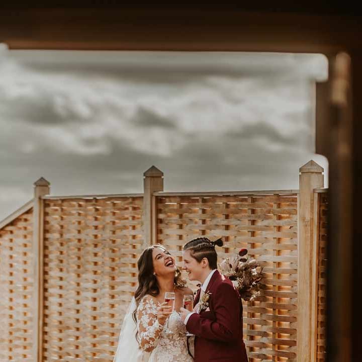 Bride in long sleeve lace illusion sleeve wedding dress with overlay drinking champagne with bride in maroon bridal suit with grey waistcoat, dusky pink tie and autumnal dried flower boutonniere 