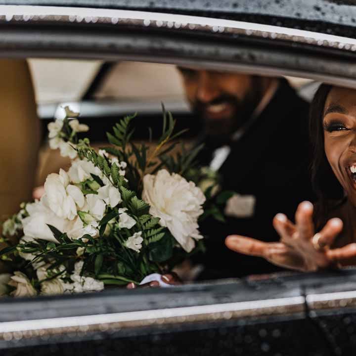 Bride holding white garden rose, white hydrangea, cows parsley and foliage bridal bouquet waving at guests from classic black wedding car 