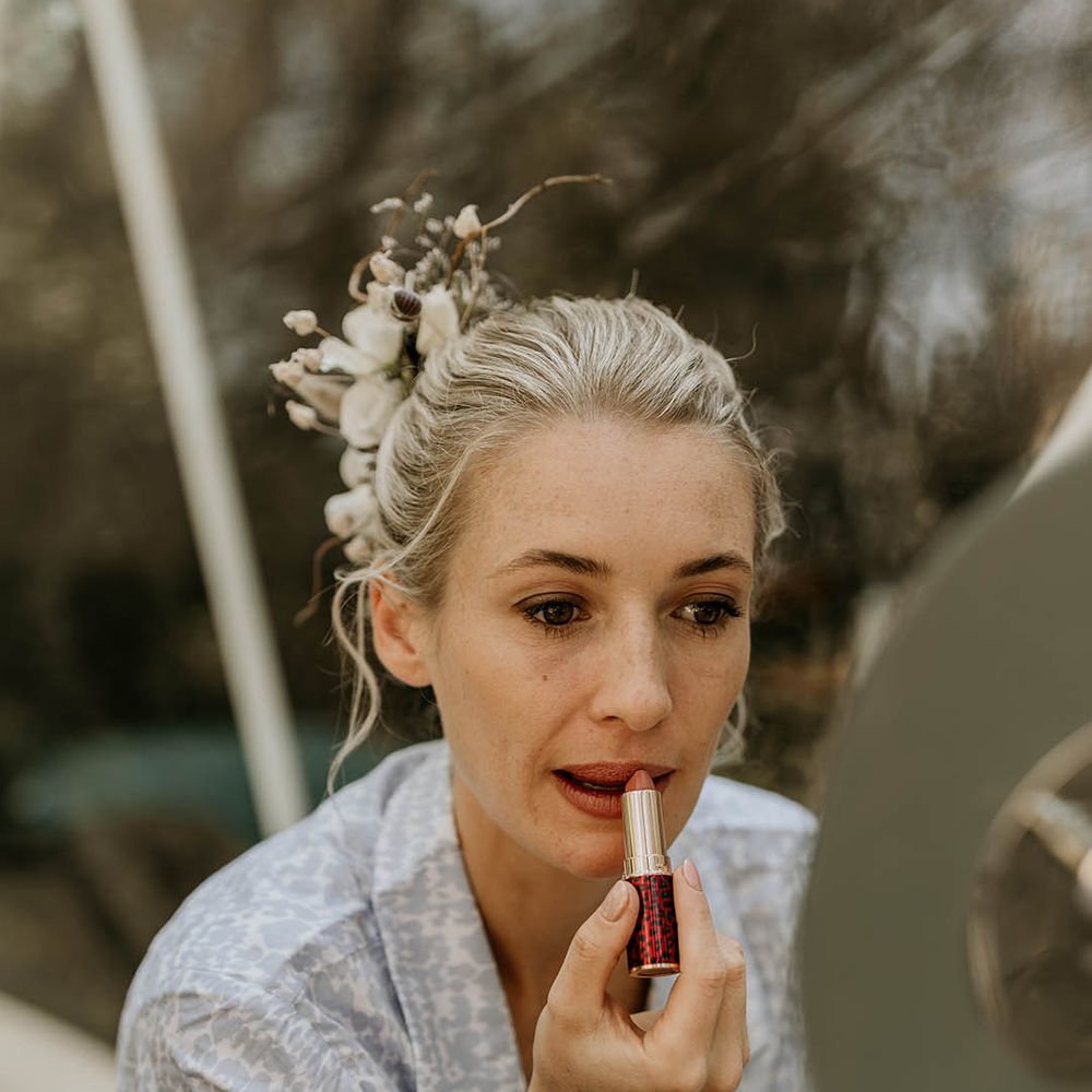 Bride in light blue and white wedding pyjamas doing her own wedding makeup with her hair in a sleek updo with white flower hair accessory 