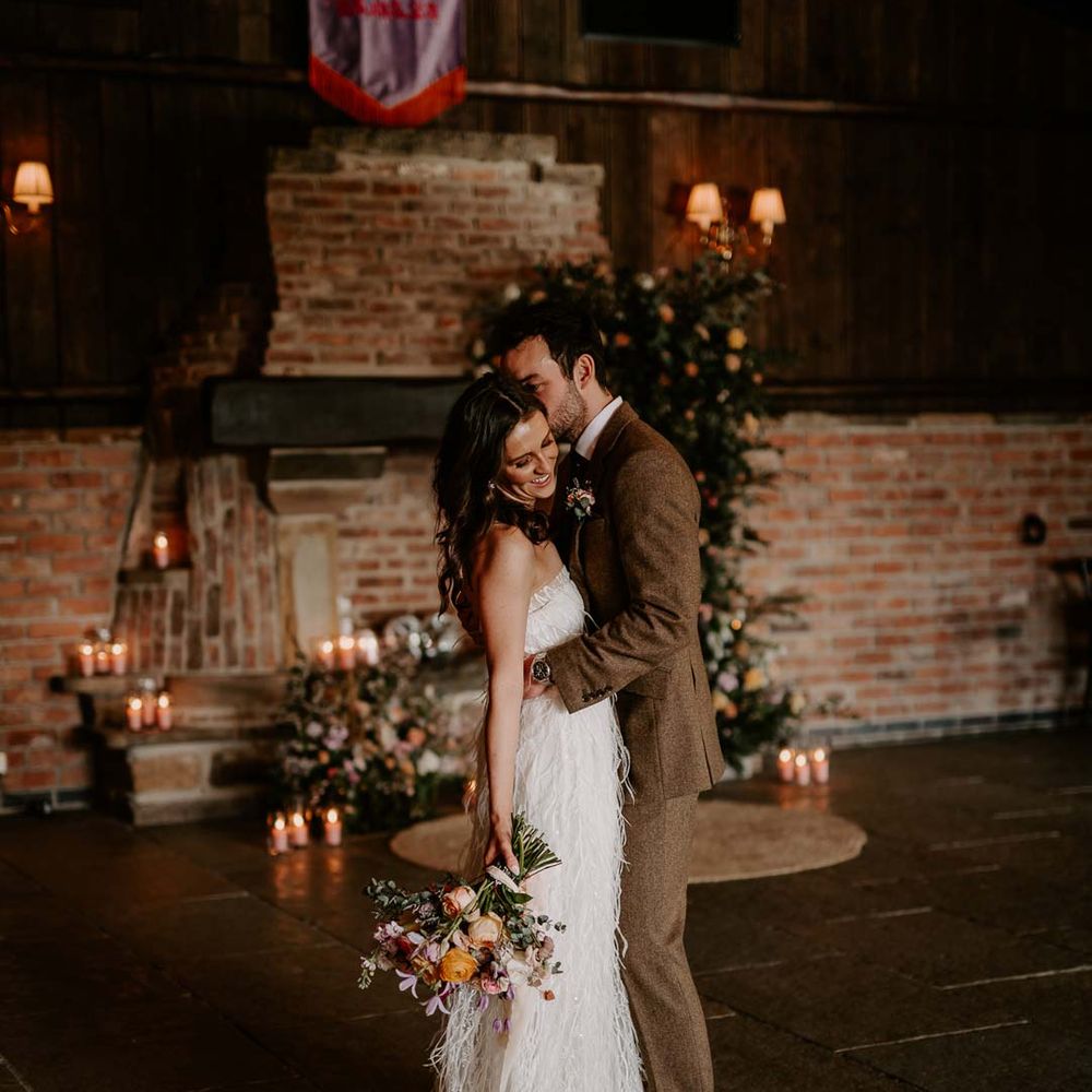 Bride in strapless ivory fringed crepe gown and chapel length veil holding mixed bridal bouquet with garden roses, carnations, peonies and foliage posing with groom in chocolate brown grooms suit in reception room of Willow Marsh Farm Loughborough 