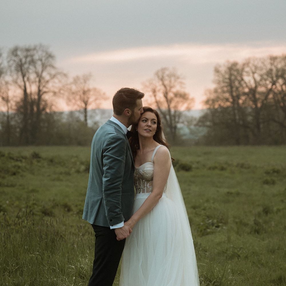 Groom in stunning blue velvet blazer kisses the bride on her forehead at sunset 