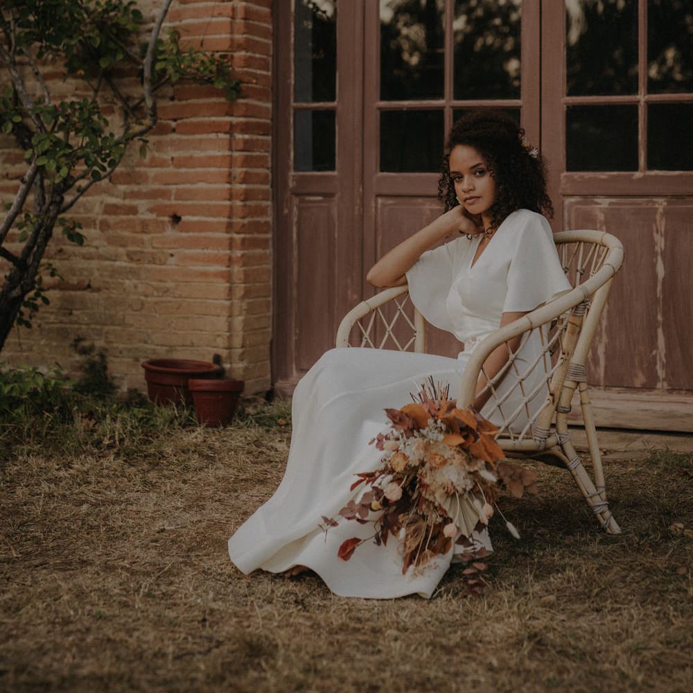 Birde in a minimalist wedding dress with floaty sleeves sitting in a wicker chair holding an orange and brown leaf and flower wedding bouquet 