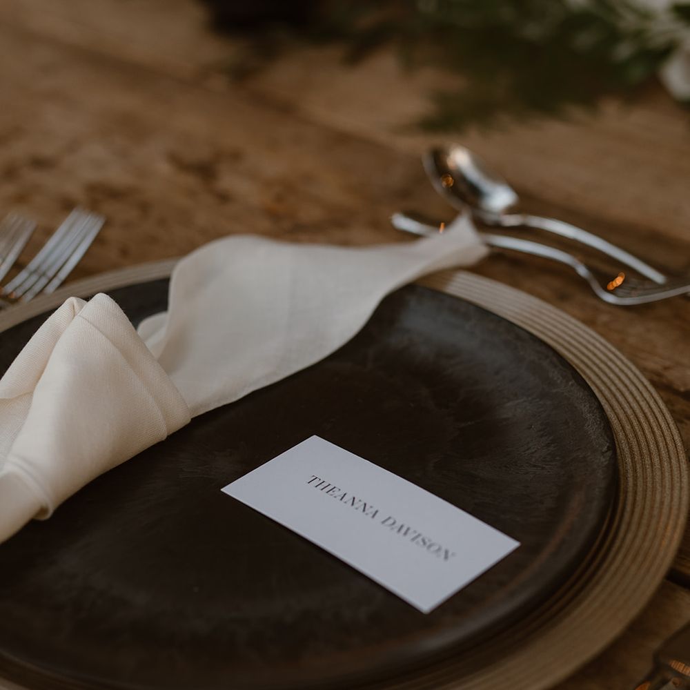 Black dinner plate with printed table name card, white napkins and silver cutlery on rustic wooden wedding table