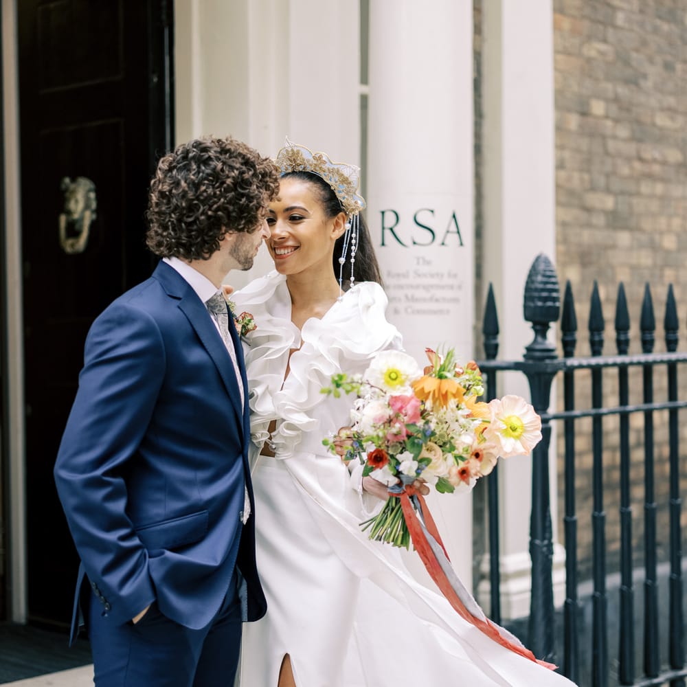 Bride & groom stand outdoors whilst looking lovingly at one another whilst bride holds floral bouquet with ruffled front