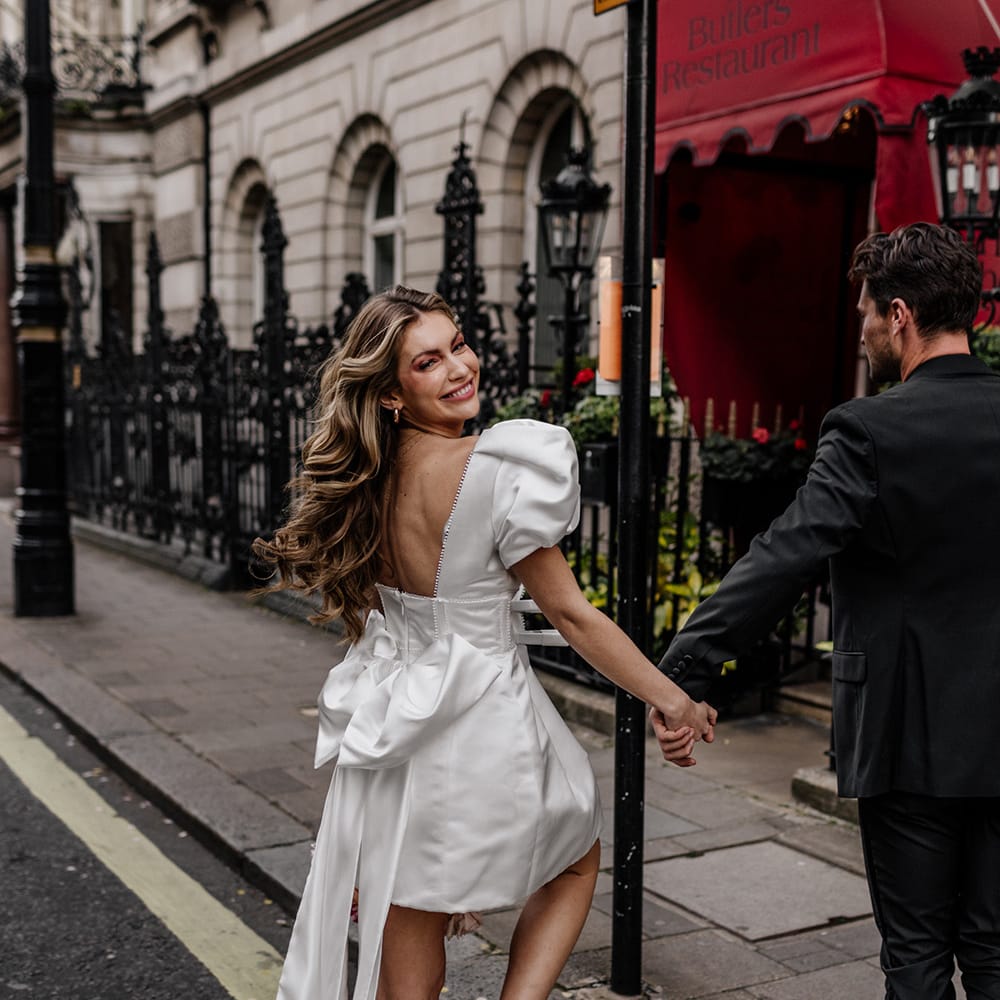 Bride with long wavy hair wearing a short wedding dress with bow back detail and pink mules, stepping onto the curb 