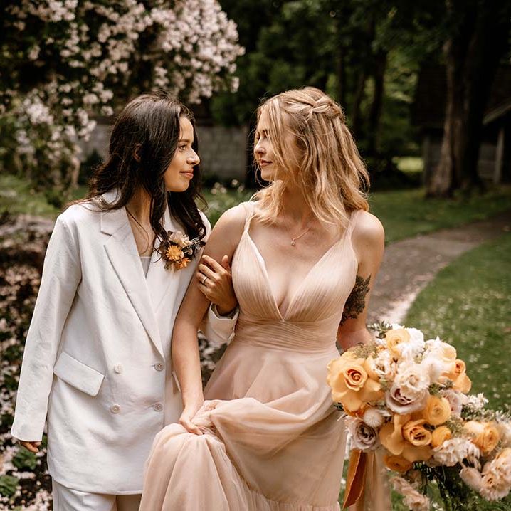 Bride in a white suit and a blush wedding dress holding a honey yellow wedding bouquet walking arm in arm 