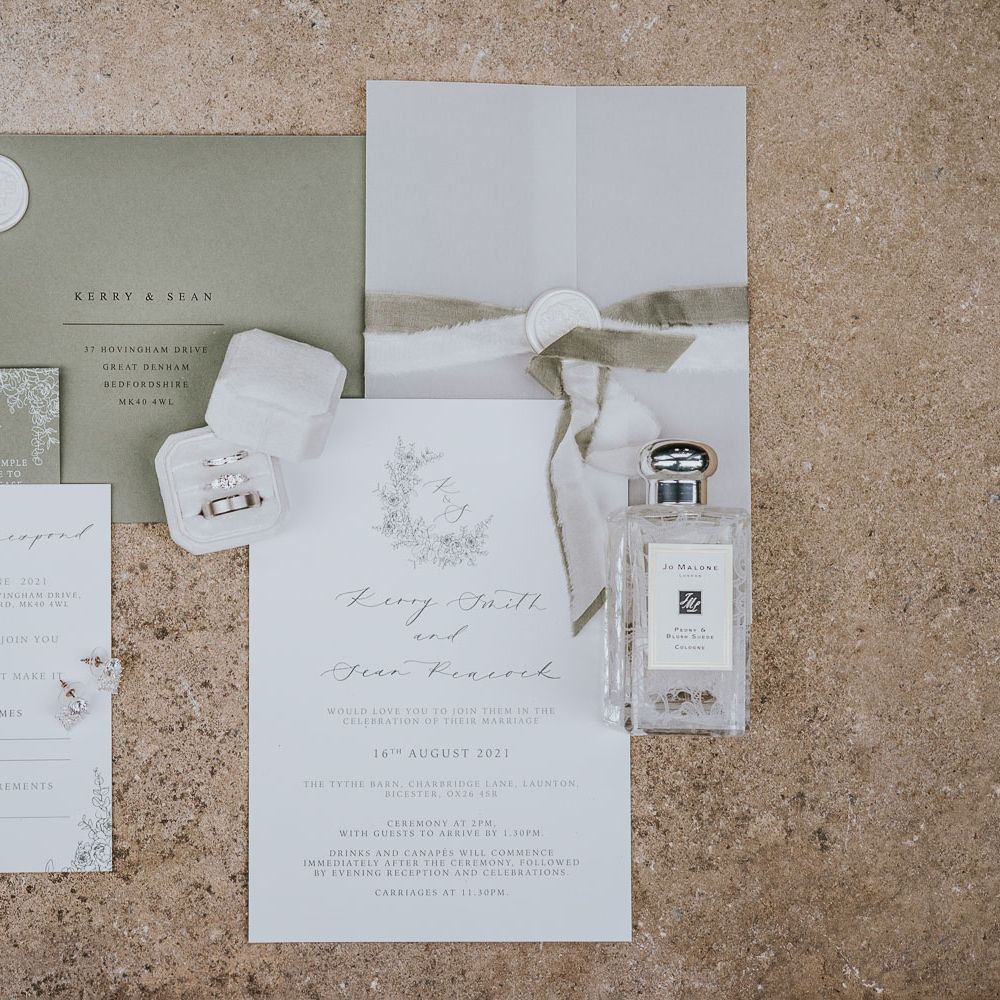 Green and white stationery with white wax seals, jo Malone perfume bottle, diamond earrings and wedding rings in white box at Tythe Barn wedding with barn wedding flowers