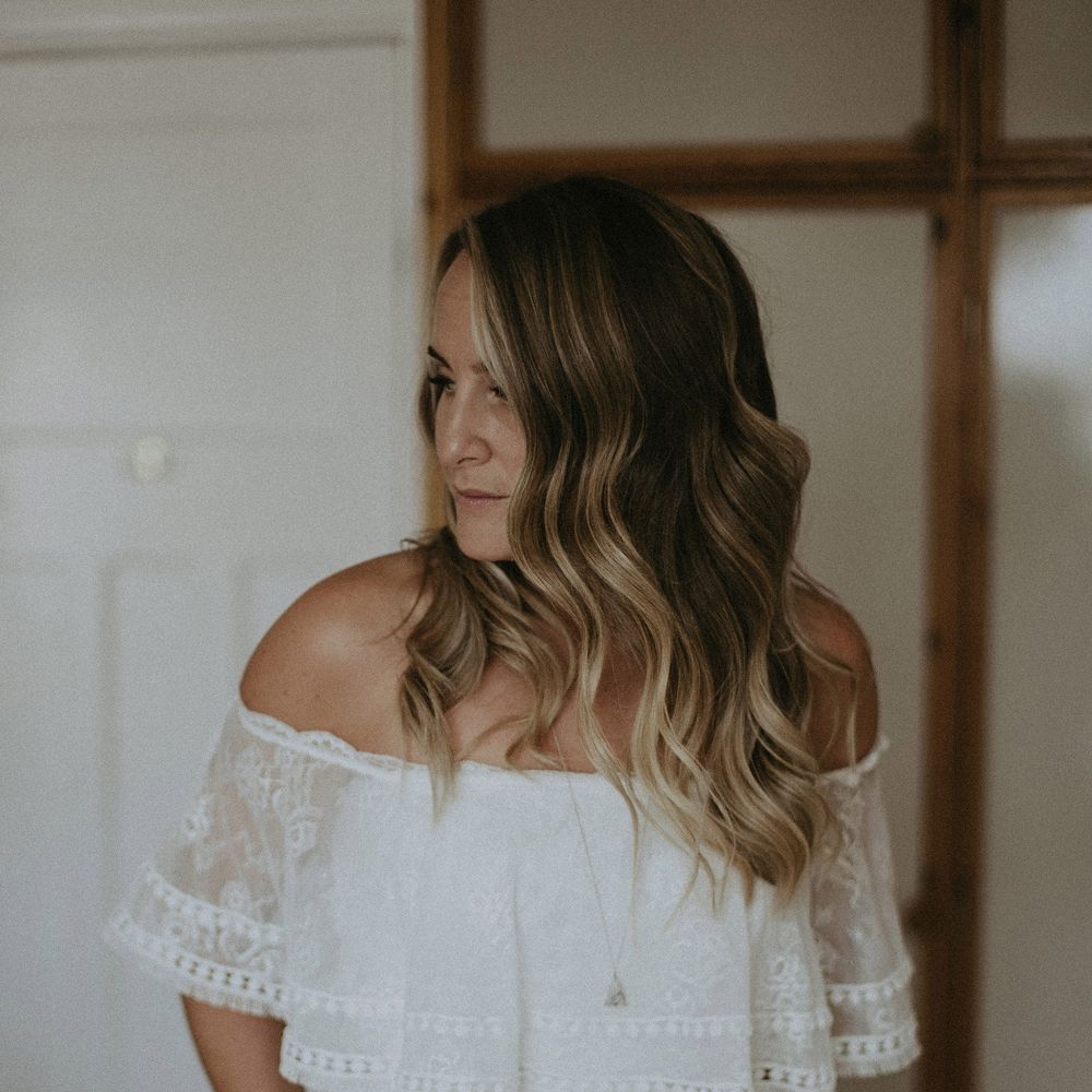 Bride with curled hair in Grace Loves Lace Bardot wedding dress and long silver necklace before Isle of Wight wedding with macrame wedding decor