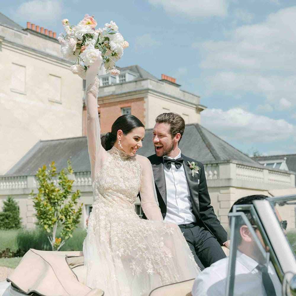 Bride in a lace long sleeve wedding dress sitting on the back of a vintage wedding car waving her bouquet int he air 