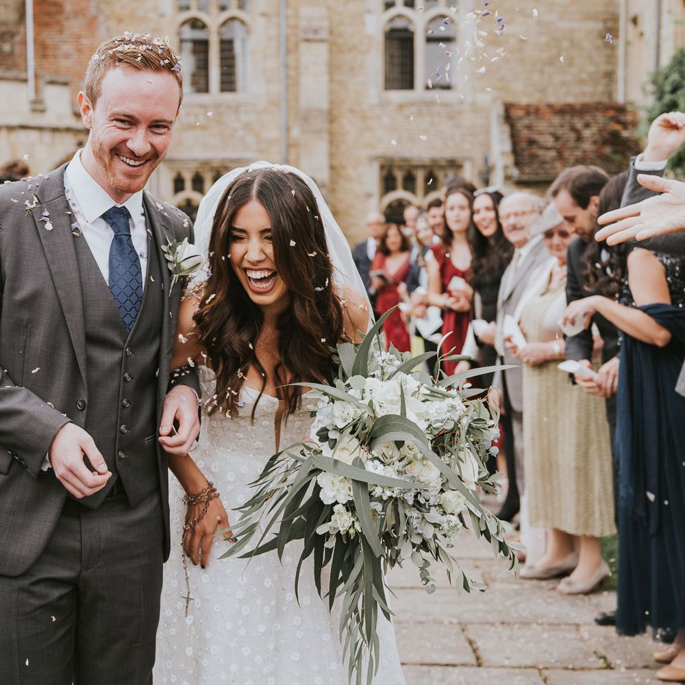Laughing bride in Pronovias wedding dress and veil holding white and green bridal bouquet walks with groom in grey three piece shot, blue tie and white rose buttonhole as guests throw confetti at Notley Abbey wedding