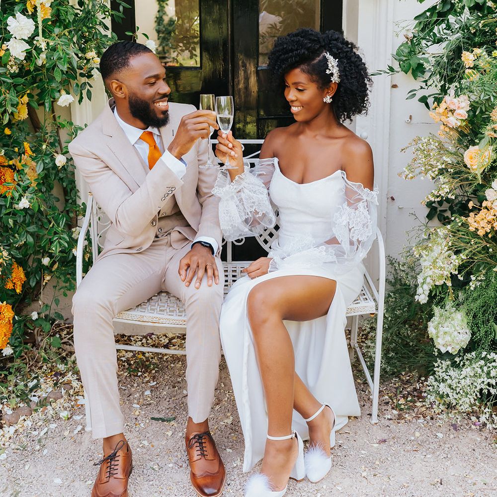 Bride in a strapless wedding dress with sheer sleeves drinking champagne with her groom in a stone coloured suit outside their Modern Hall wedding venue 