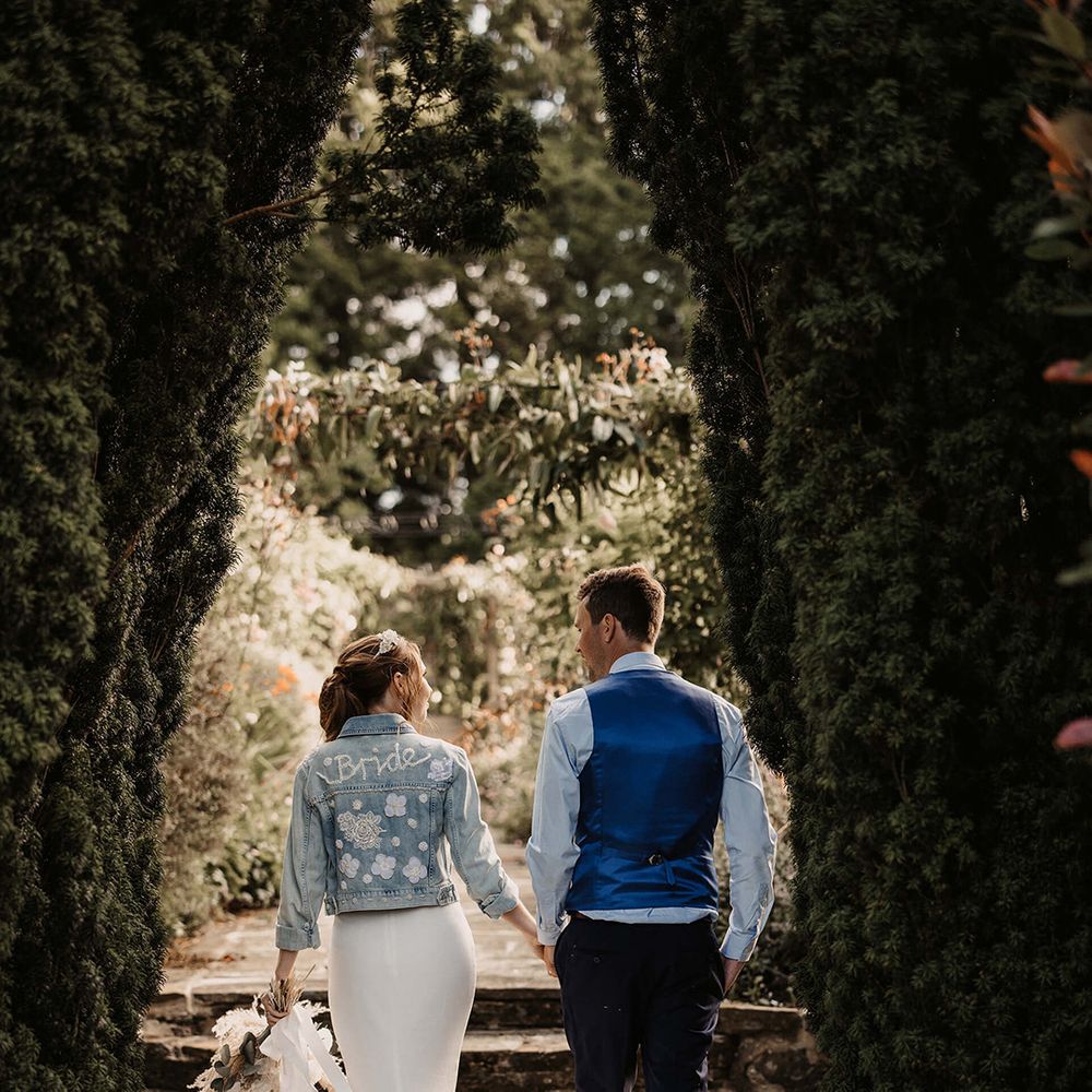 Bride & groom walk hand in hand and bride wears hand stitched denim jacket