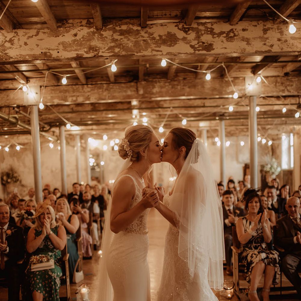 Industrial wedding with two brides in a lace and fishtail wedding dress kissing at the altar under a canopy of festoon lights 