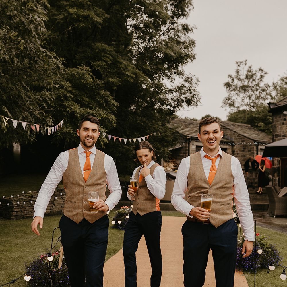 Groomsman in brown waistcoats and orange ties make funny faces and smile at the camera together