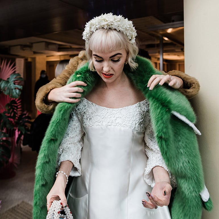 Bride with white flower headband and pearl handbag gets help putting on her green fur coat 
