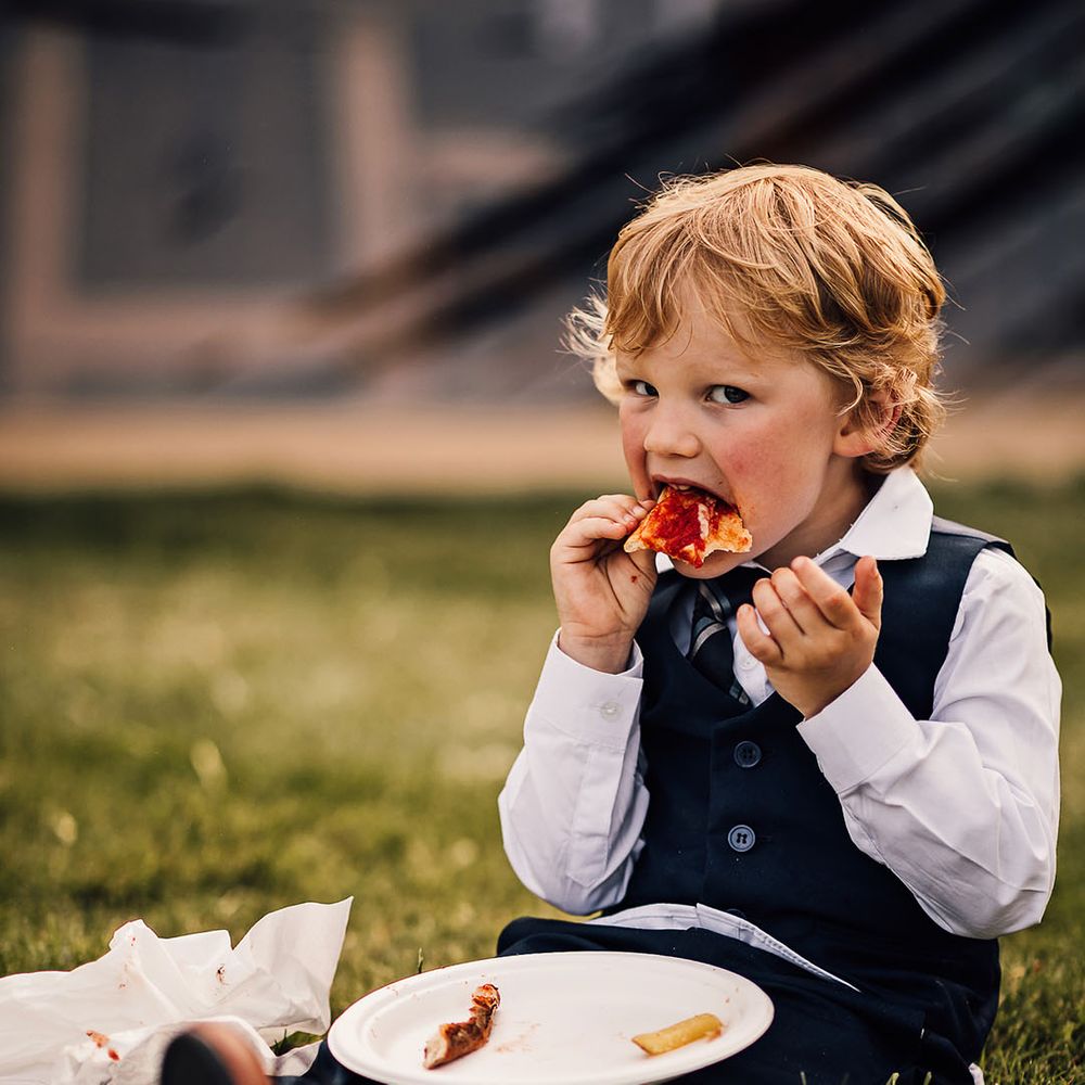 Little boy wedding guest eats a slice of pizza on a paper plate