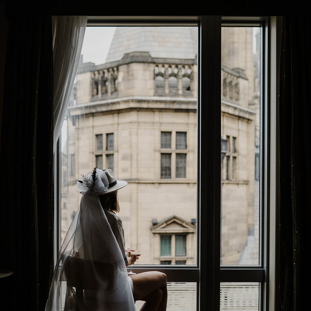 Bride wearing fedora hat with veil attached looking out of the window 