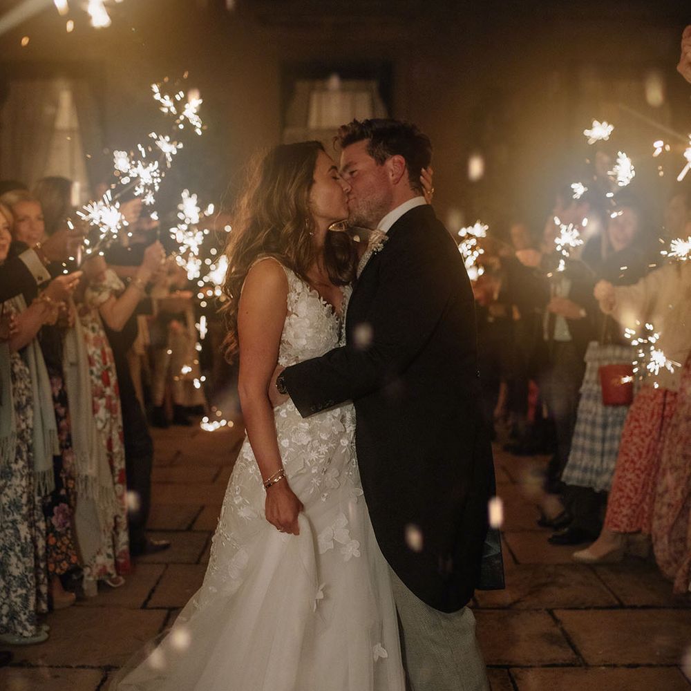 Bride in Mira Zwellinger wedding dress kisses groom in morning suit as guests wave sparklers at the end of the night 