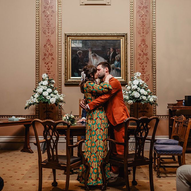Bride wearing brightly coloured La Double J wedding gown & groom in orange Paul Smith suit kiss at Bristol Register Office 
