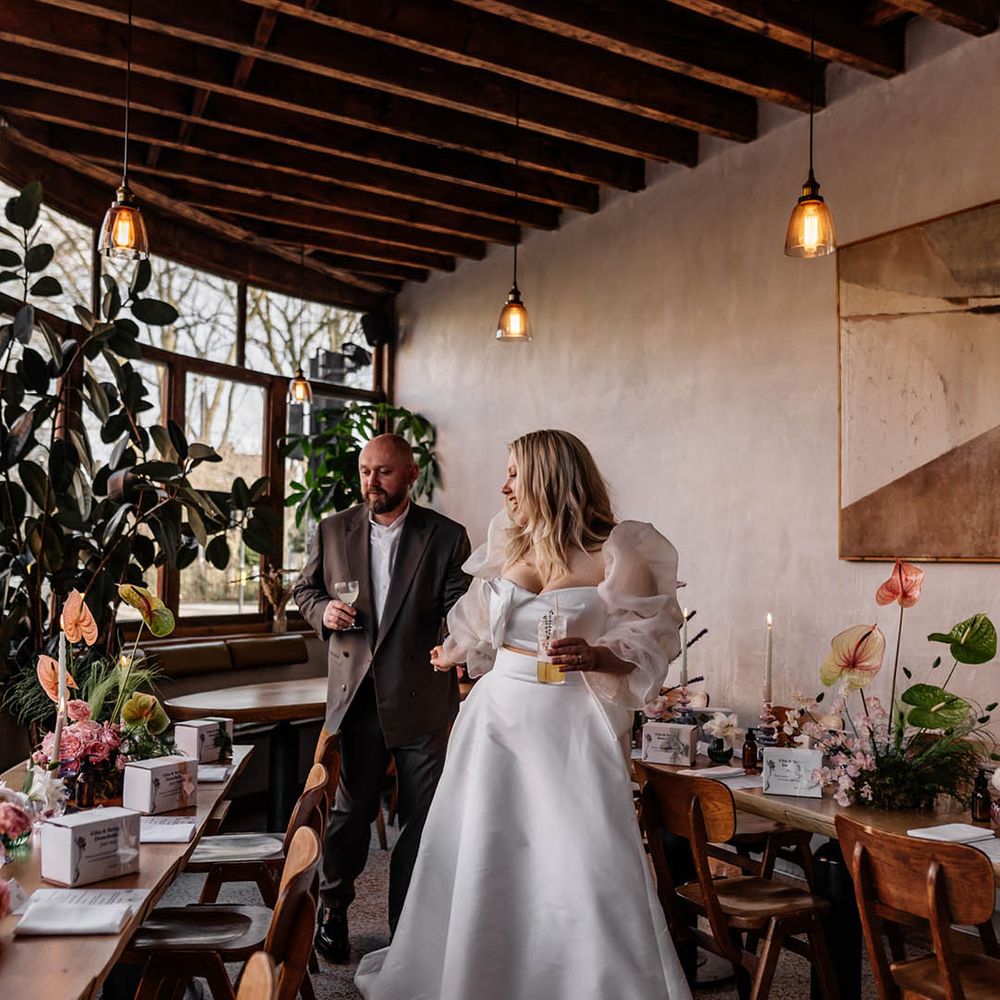 Bride in a Jesus peiro wedding dress holding hands with her groom in a double breasted jacket looking at their wedding reception table decor, flowers and favours 