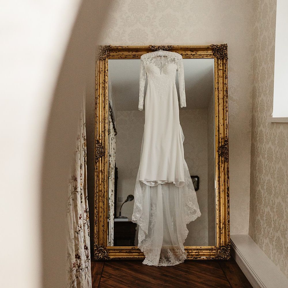 Lace bridal gown with long sleeves hangs in front of gold mirror 