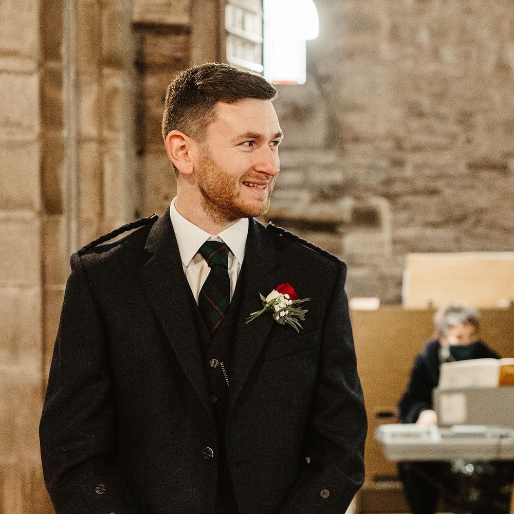 Groom awaits his bride wearing three piece suit complete with kilt and white and red floral buttonhole 