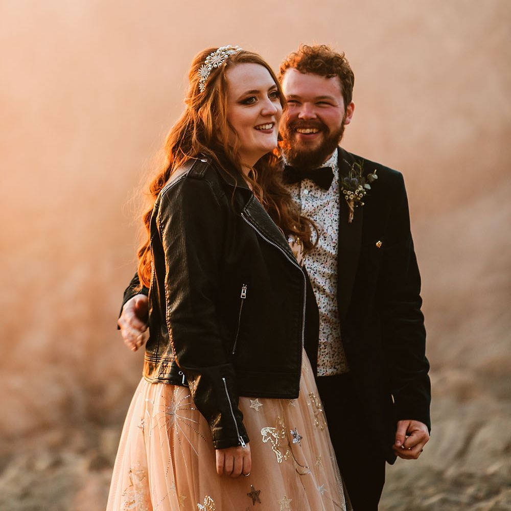 Bride wears personalised leather jacket and pearl embellished wedding crown whilst stood with her groom within orange smoke bomb