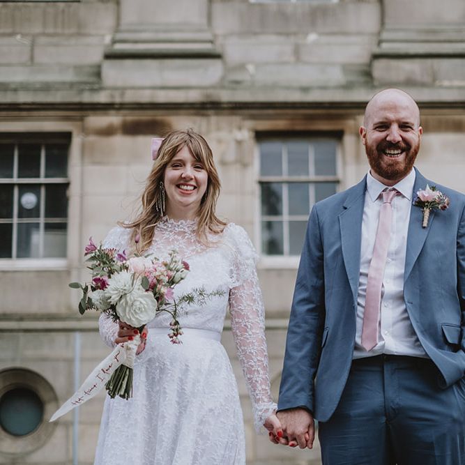 Bride wears high neck lace vintage wedding dress whilst holding hands with her groom who wears high street suit with pale pink silk tie