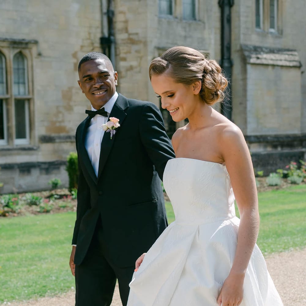 Groom in black tie wearing a colourful buttonhole walks with the bride in a Jesus Peiro brocade wedding dress 