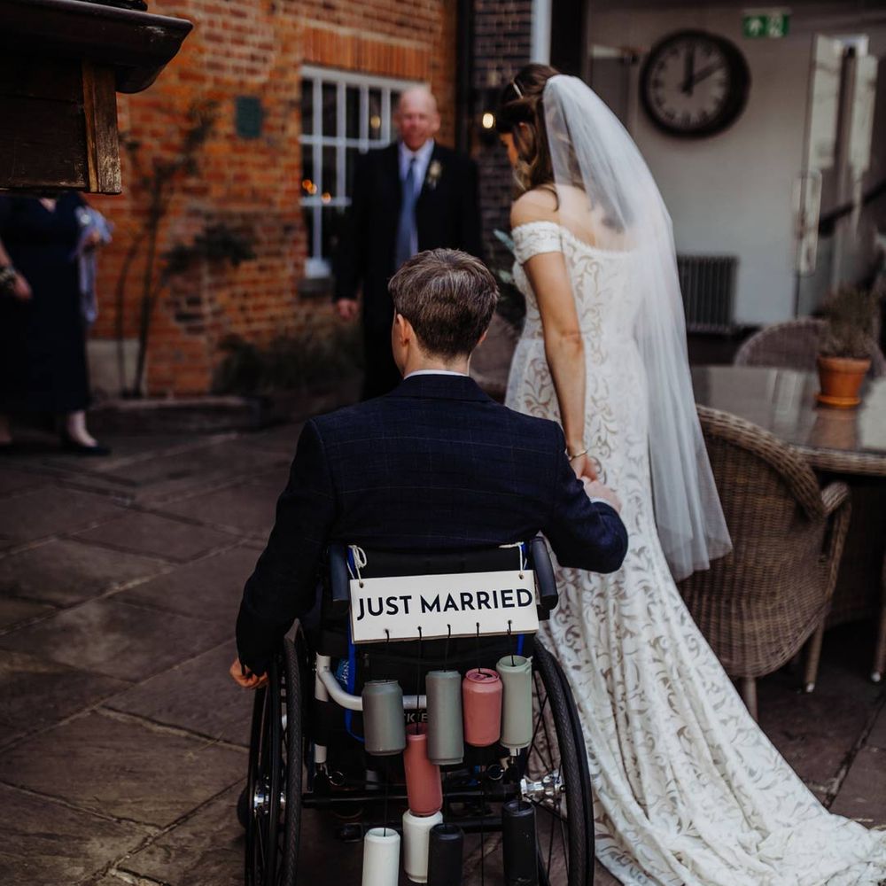 Bride in lace off shoulder dress with veil and groom in dark suit in wheelchair with DIY Just Married sign and colourful painted tin cans decorations