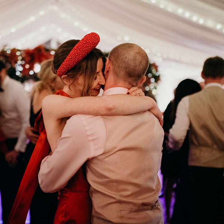 Bride in red silk wedding dress with cape, large red embellished head band and groom in waistcoat and white shirt embracing and dancing at winter wedding party