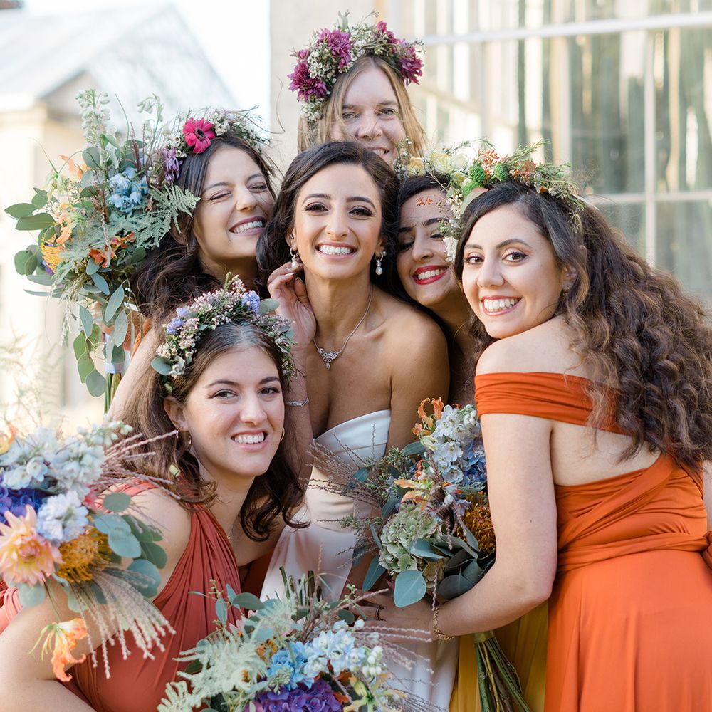 Bride stands with her bridesmaids who wear colourful ombre styled bridesmaid dresses in orange and yellow complete with floral crowns