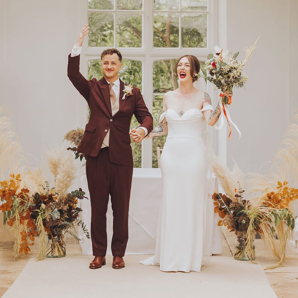 Bride holds up Autumnal styled bouquet and walks alongside her groom who wears maroon suit complete with floral buttonhole