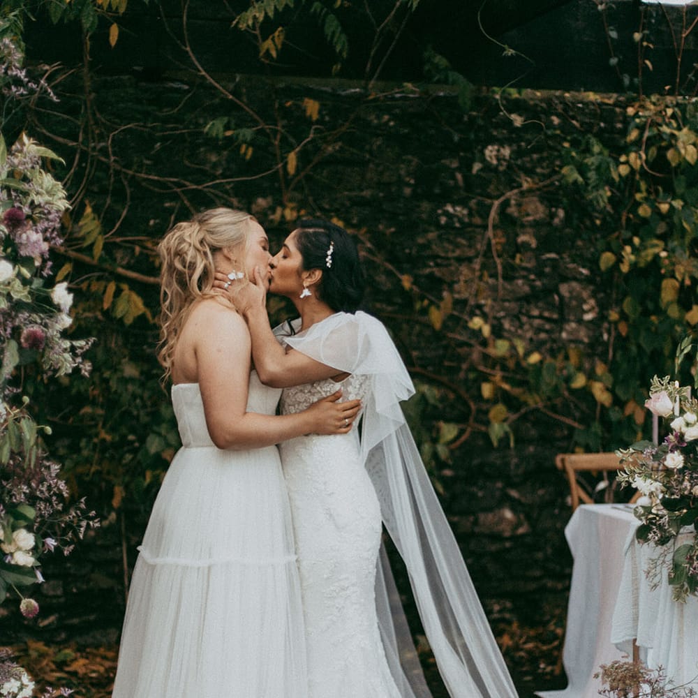 Brides in complimenting lace wedding dresses, one with short puff sleeves and the other strapless, kissing at the alter