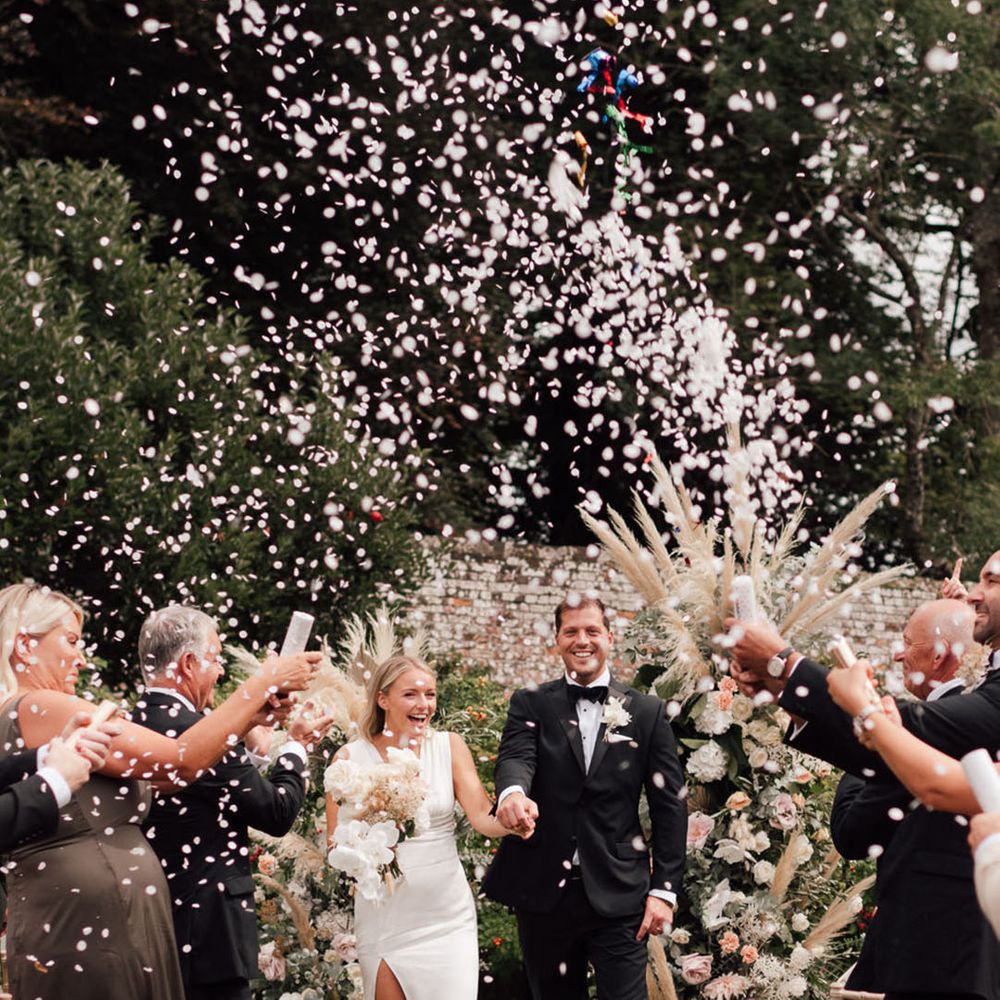 Bride and groom walk back down the aisle as white confetti explodes over them at their outdoor ceremony 