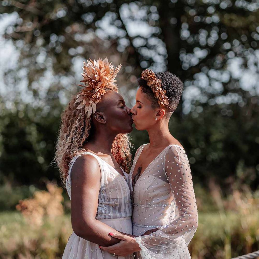 Bride in crystal embellished gown and gold headdress kissing bride in sleeveless floaty peach gown and golden leaf crown  
