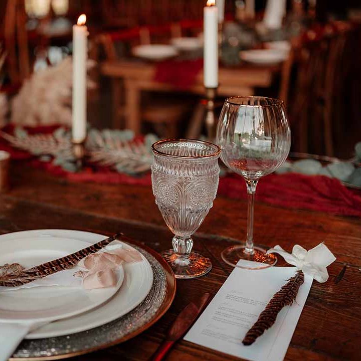 Autumnal, boho wedding tablescape; vintage wine glasses with gold rims, hexagonal golden vase, gold candle sticks with white tapered candles, foliage and pampas grass decor and pheasant feather decor