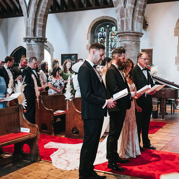 Groom, groomsmen, father of the bride and bride stand with order of service booklets listening to the officiant for the wedding ceremony 